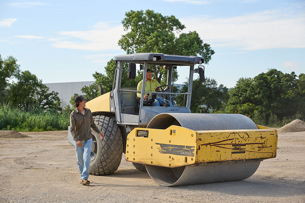 A Dreamworx employee drives a yellow Atlas Copco CA2500 soil compactor over a dirt lot, with dust rising around the large front roller.